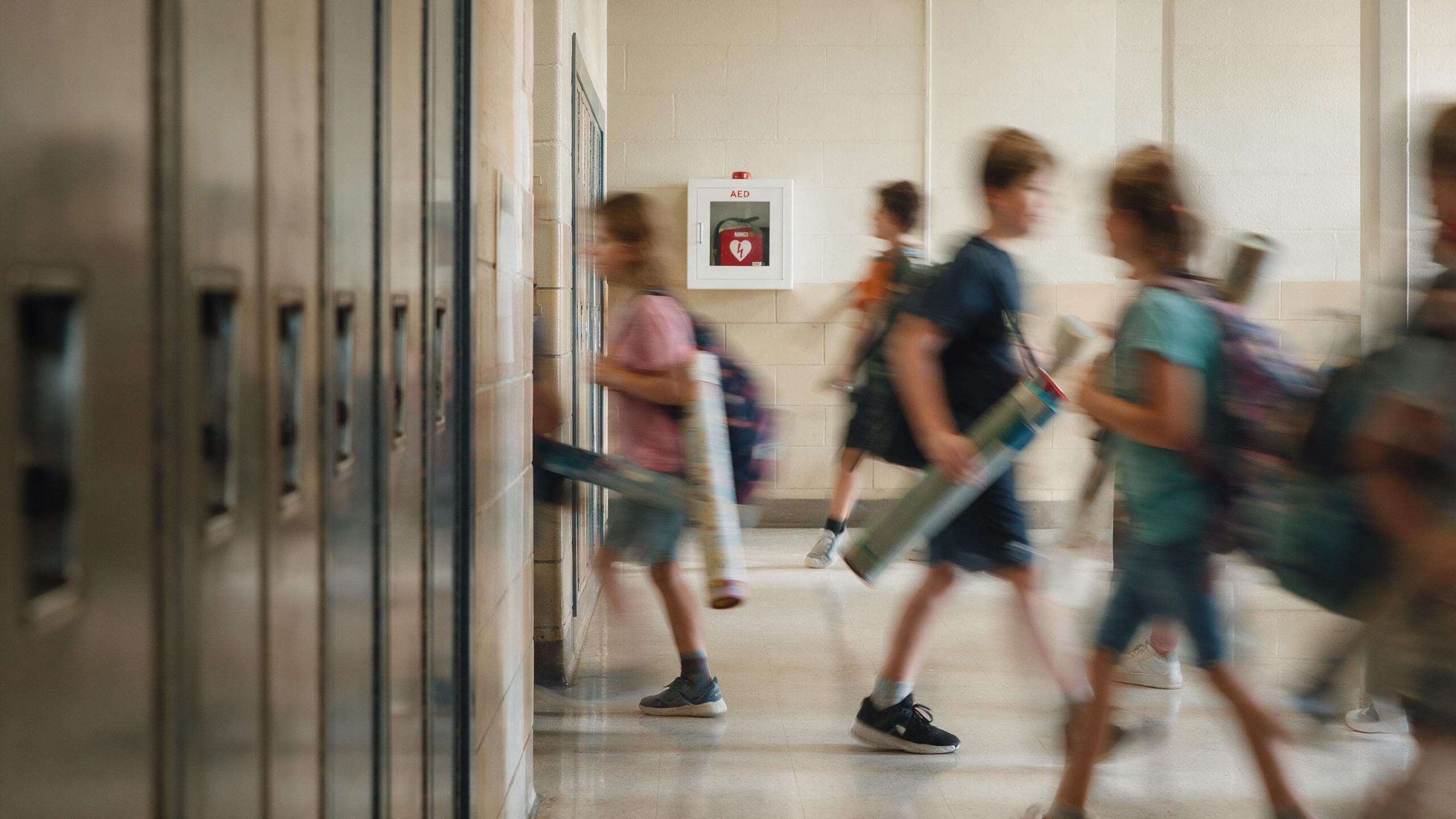 Students walking down a school hallway past a wall-mounted AED cabinet
